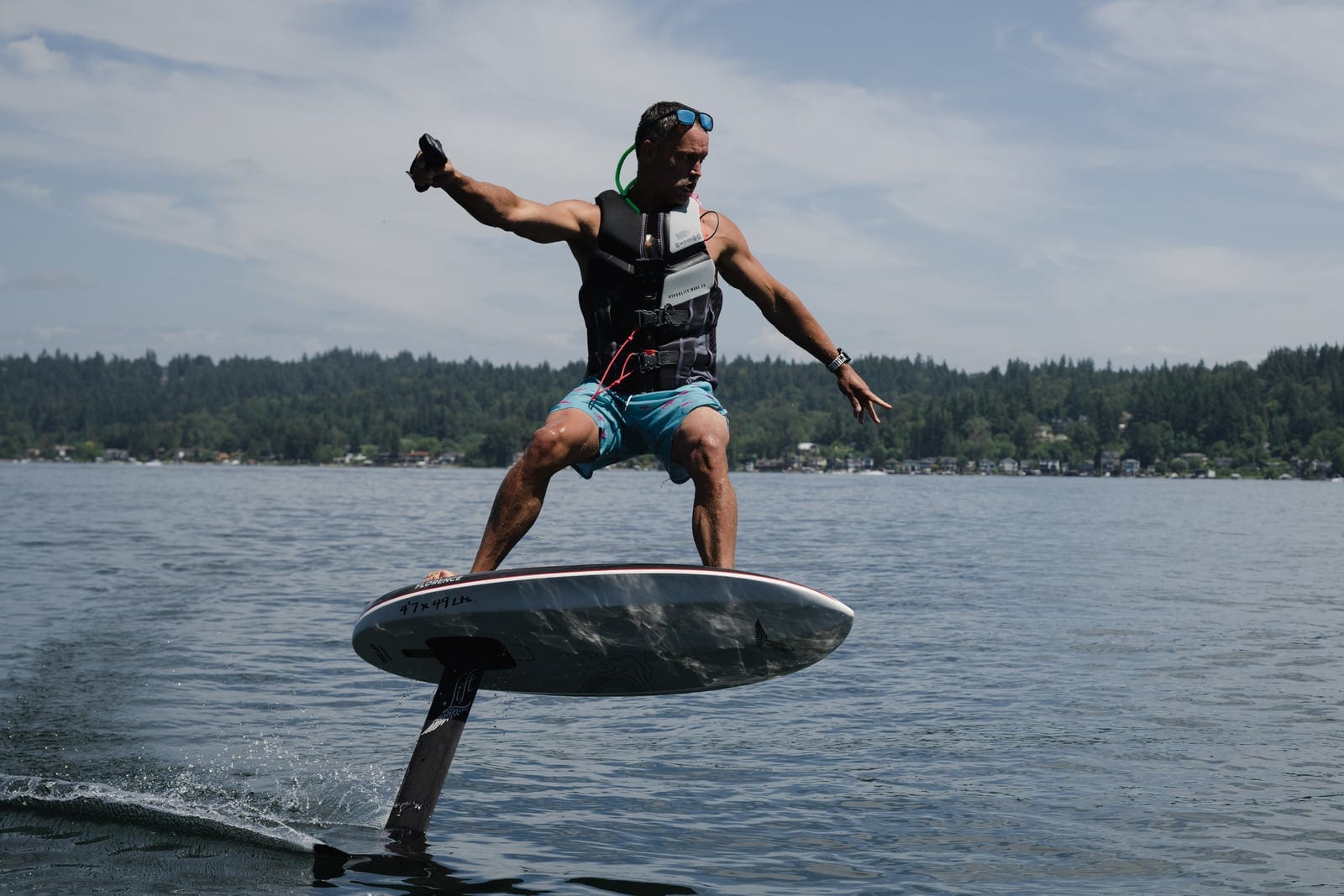 A rider carving an eFoil board clear of the water on a forested lake, wearing a Garmin watch on the left wrist.
